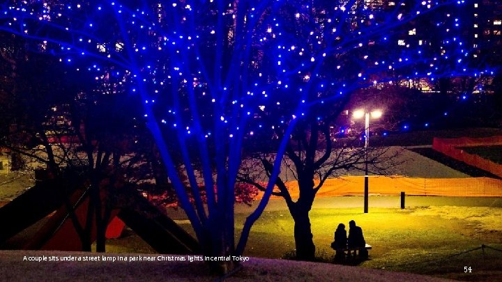 A couple sits under a street lamp in a park near Christmas lights in