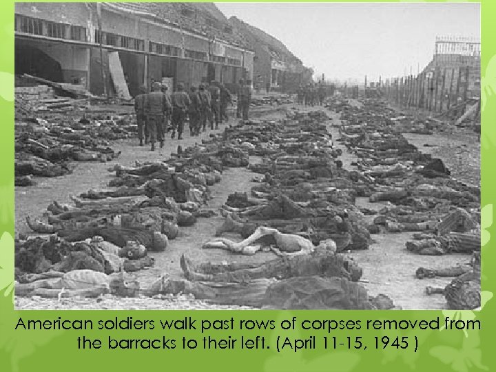 American soldiers walk past rows of corpses removed from the barracks to their left.